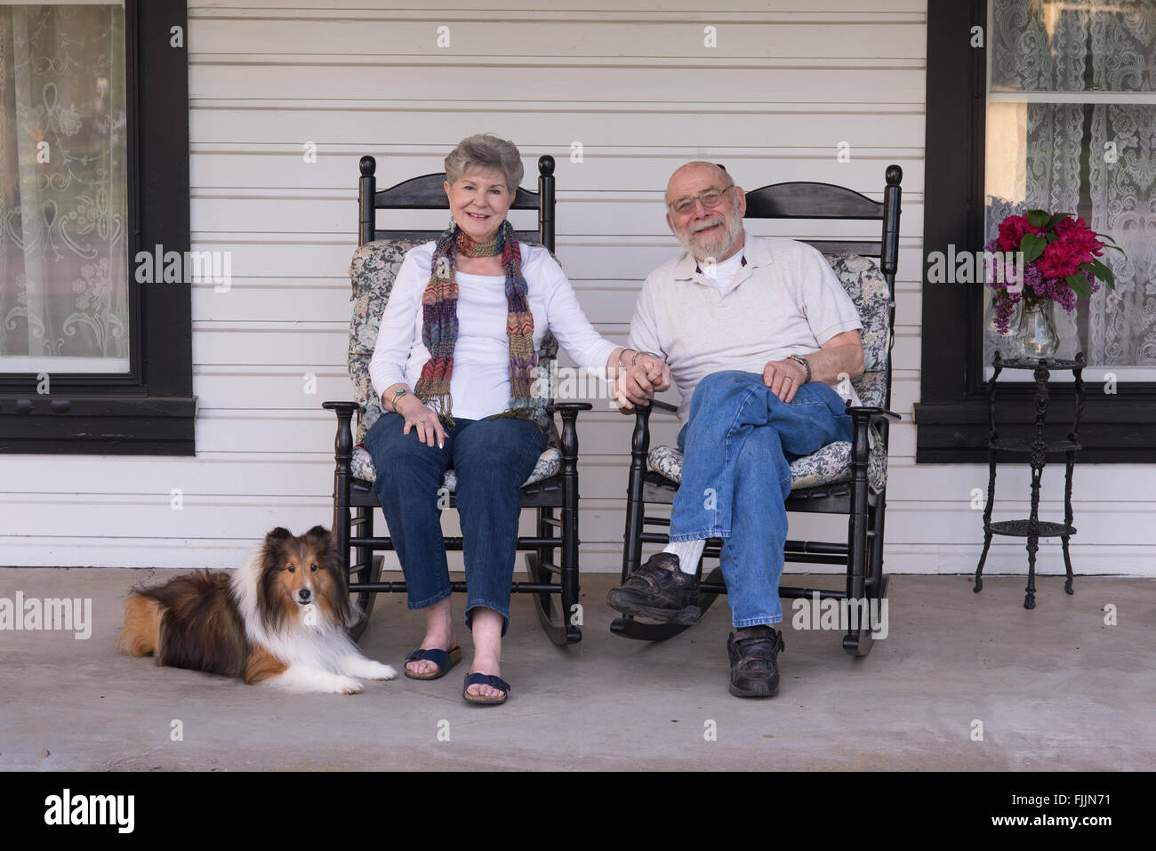 A happy married couple relax on their porch in matching rocking chairs with their Shetland sheepdog by their side. Stock Photo