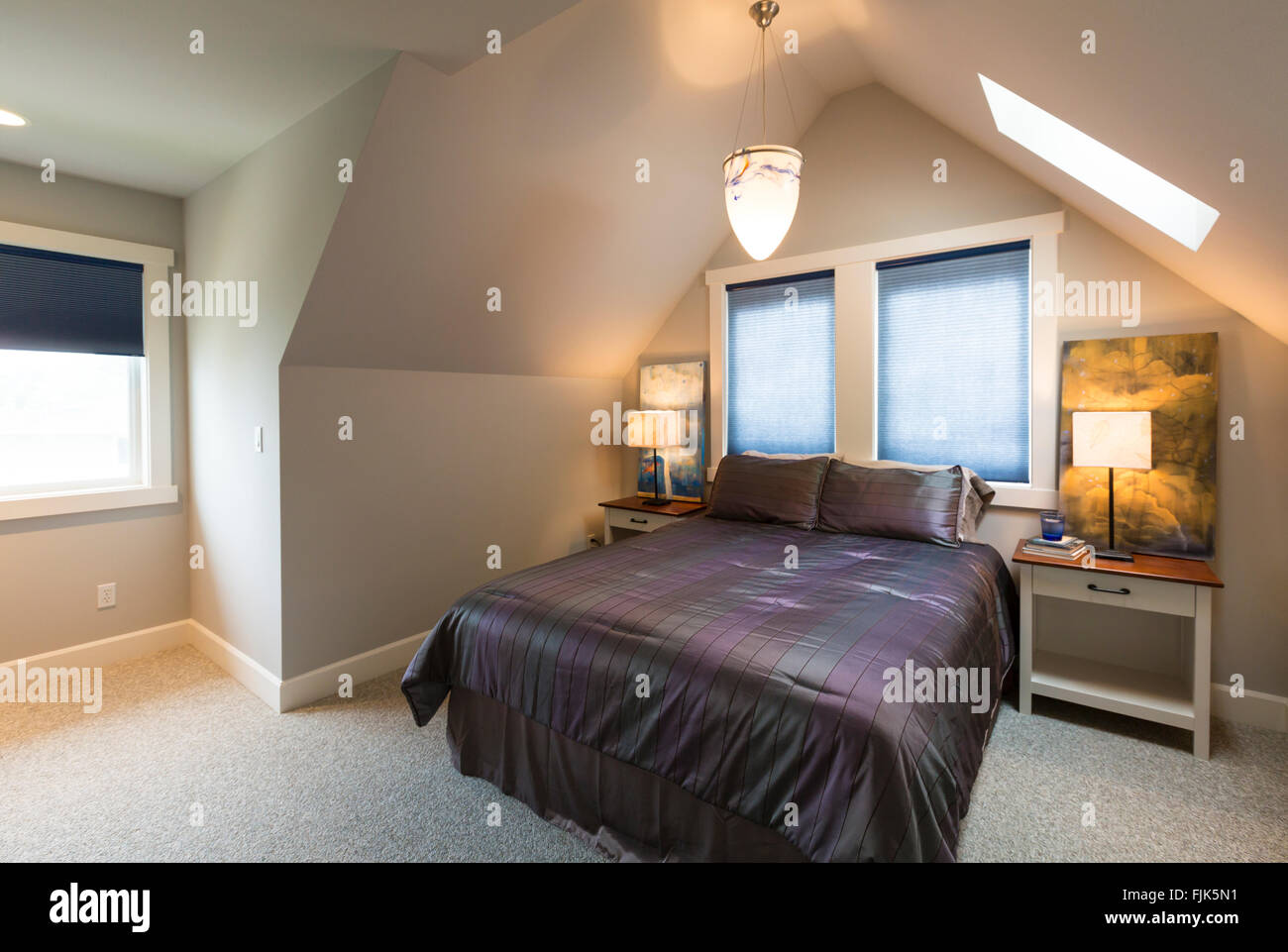 Bedroom with bed, bedside tables, vaulted ceiling, windows with window coverings, artwork and accent lighting in upscale home Stock Photo