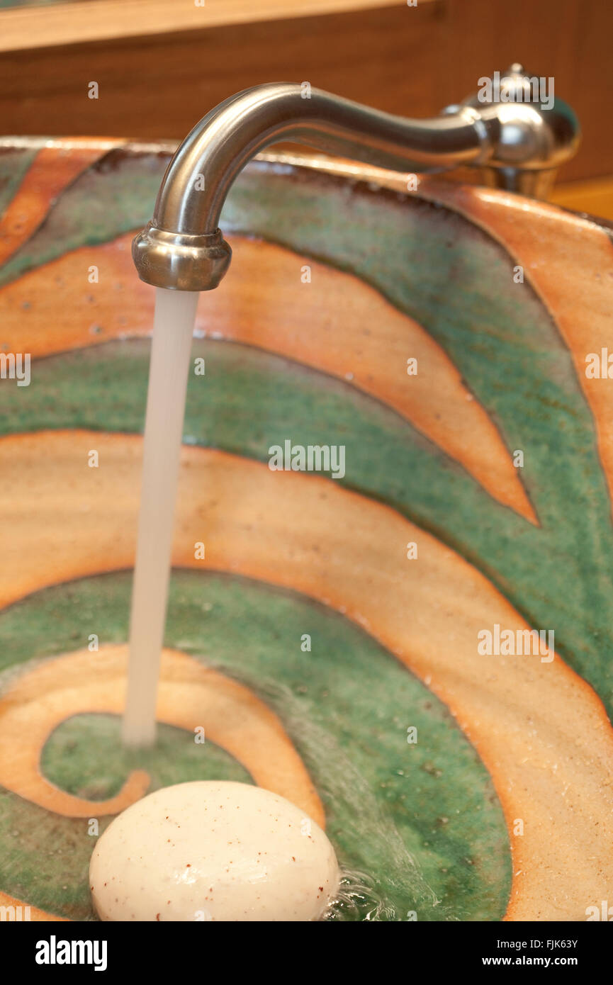 Water running from faucet tap into a creative handmade ceramic sink with a bar of soap. Washing up in creative home bathroom interior. Stock Photo