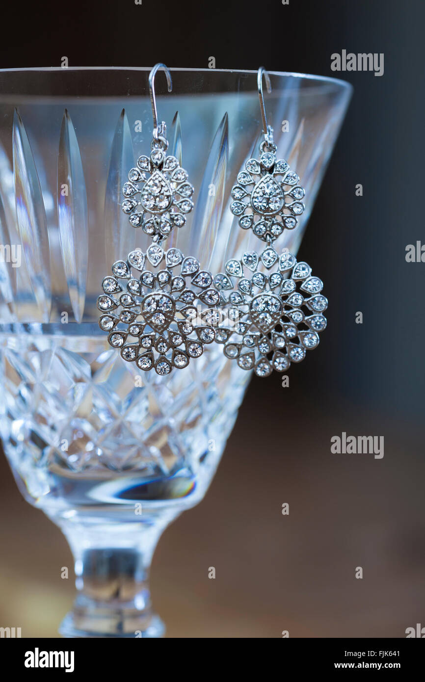 Fancy, ornate rhinestone diamond earrings hanging from the lip of a fine cut lead crystal wine glass. Expensive, valuable luxury goods. Stock Photo