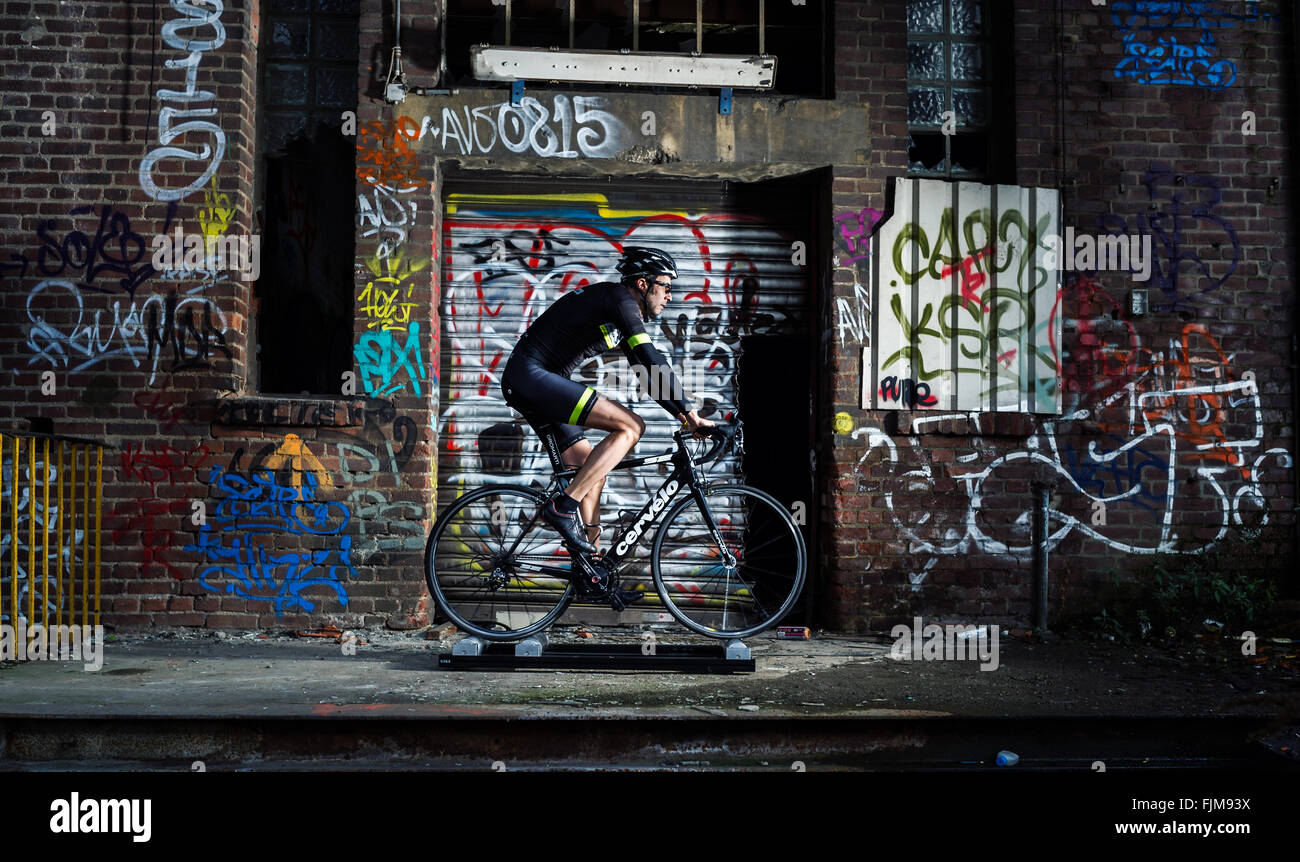 Cyclist riding on a roll trainer in front of a wrecked buling painted with graffiti. Stock Photo