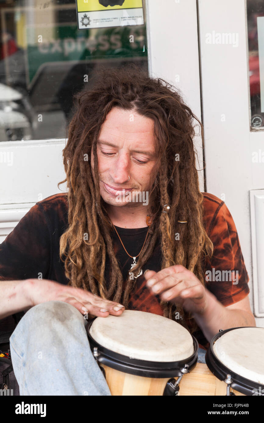 A young Caucasian Male Busker with long dreadlocks sitting on the pavement playing bongo drum outside a shop doorway Llandudno Stock Photo