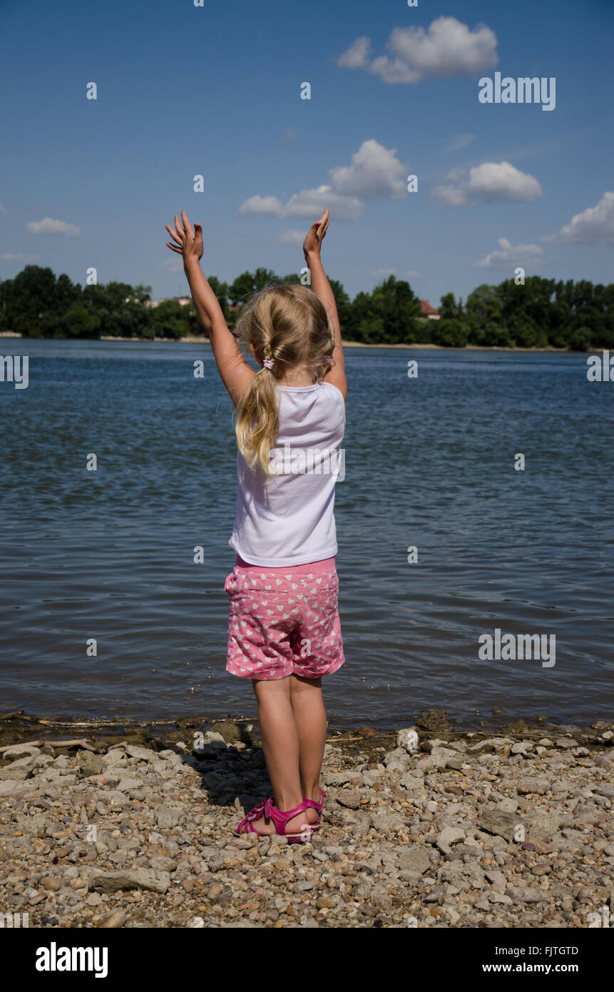 little girl with long blond hair standing in the strand by the blue river with hands up Stock Photo