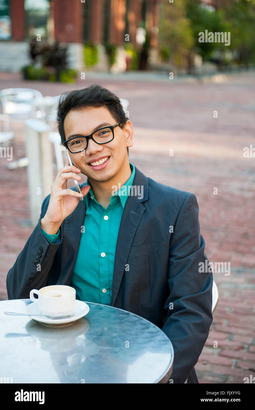 Successful young asian man in business casual attire sitting and smiling in relaxing outdoor cafe with cup of coffee talking on Stock Photo