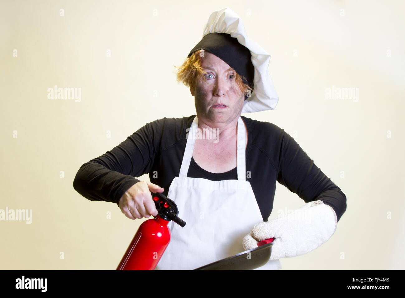 Mature woman with singed face holding pan and fire extinguisher with chefs hat and apron. Stock Photo