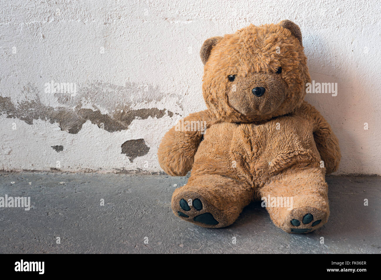 A old and dirty Teddy bear is sitting against a white concrete wall Stock Photo