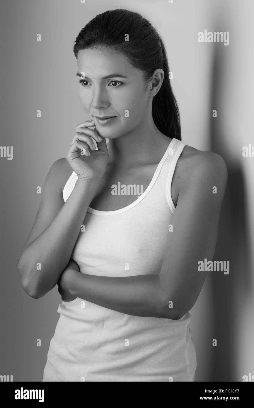 Single beautiful woman in sleeveless blouse with folded arms and hand on chin near wall in black and white Stock Photo