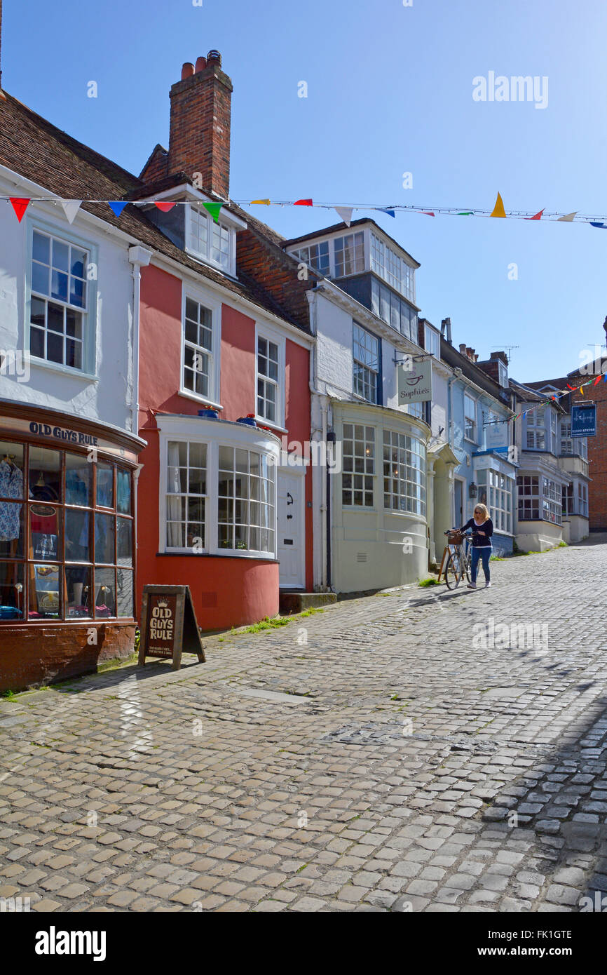 Lymington Hampshire colorful rendered walls and bay windows on shop fronts along hilly cobbled Quay Hill street woman walking with bike England UK Stock Photo