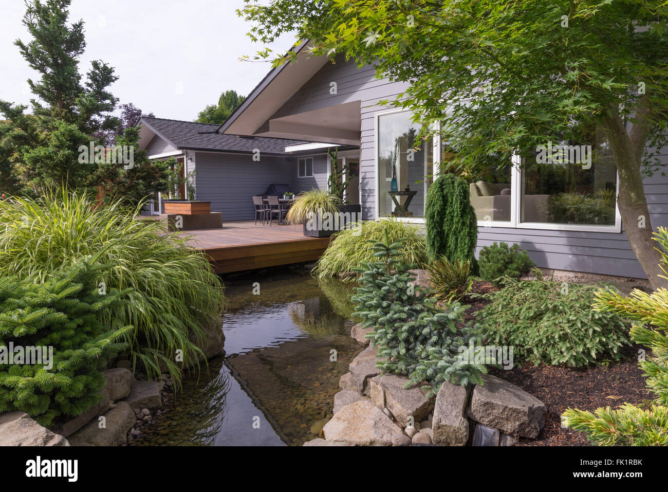 A water feature emulating a creek flows under a deck and between landscaped rocks with a contemporary home in the background. Stock Photo
