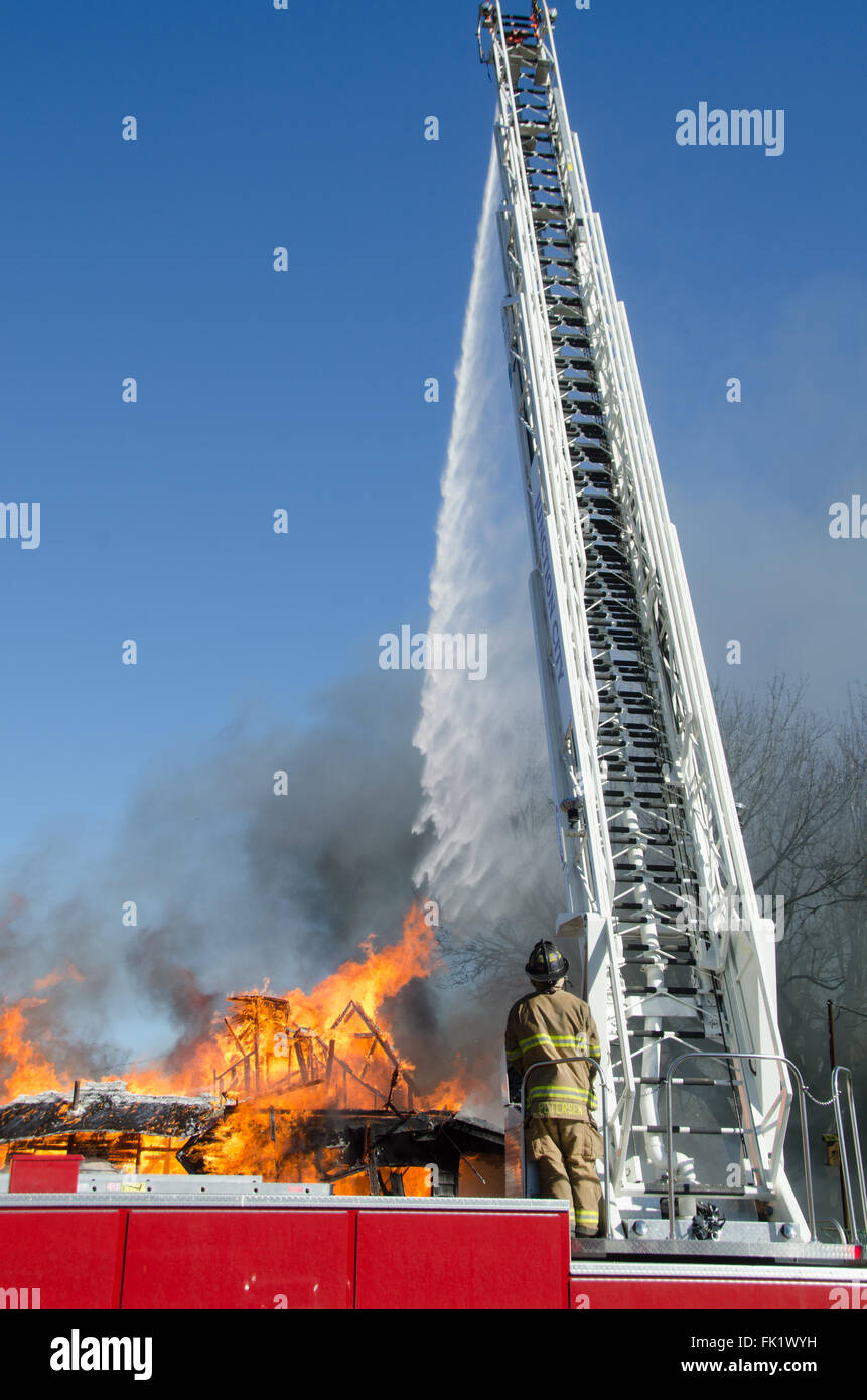 A fireman standing at the controls of an105 foot reach aerial ladder/truck directs a sream of water on a blazing house. Stock Photo