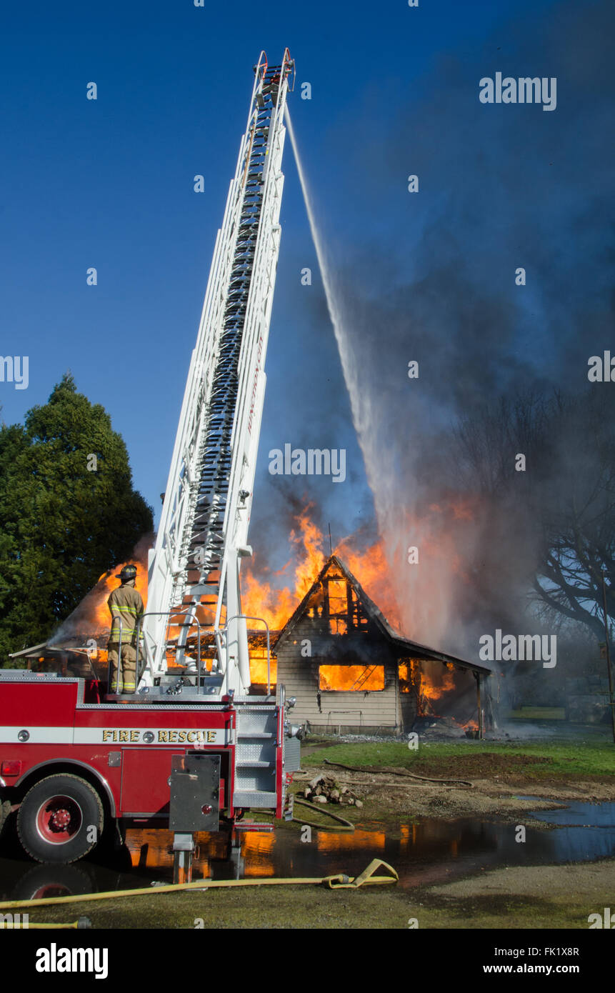 A fireman uses controls at the base of a ladder truck to douse a blazing house. Stock Photo