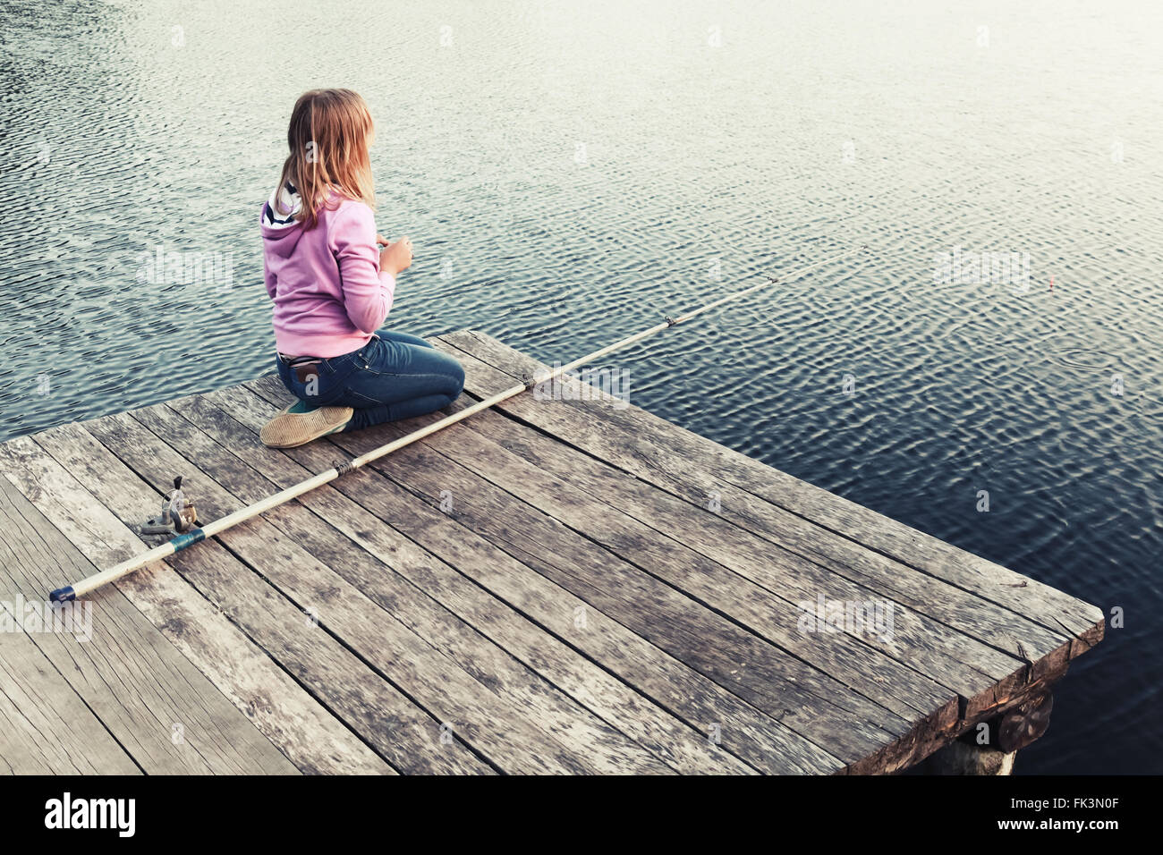 Little blond Caucasian girl sitting on a wooden pier with fishing rod ...
