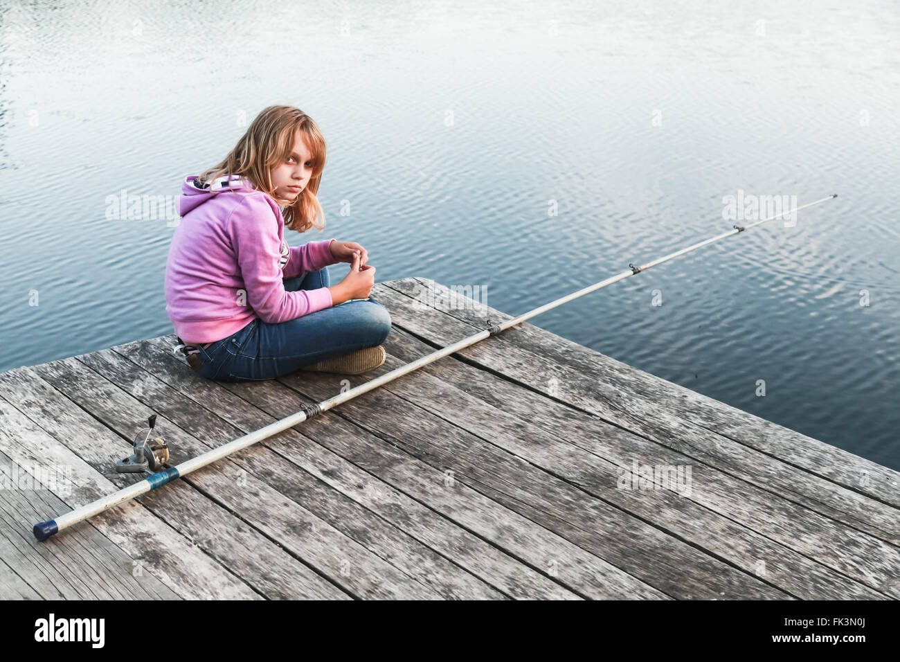 Blond Caucasian girl sitting on a wooden pier with fishing rod Stock Photo