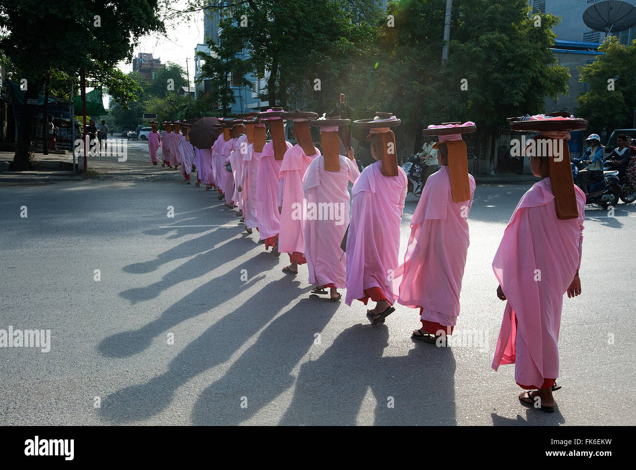 Long line of Buddhist nuns dressed in pink robes, heads covered, alms bowls on their heads, crossing main road in Mandalay Stock Photo