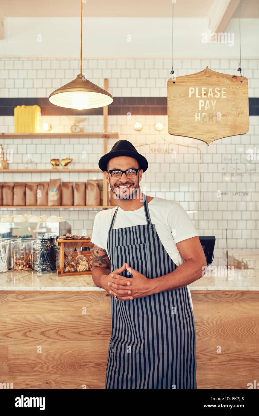 Coffee shop worker smiling to camera, standing at the counter. Happy young man in apron and hat leaning to cafe counter. Stock Photo