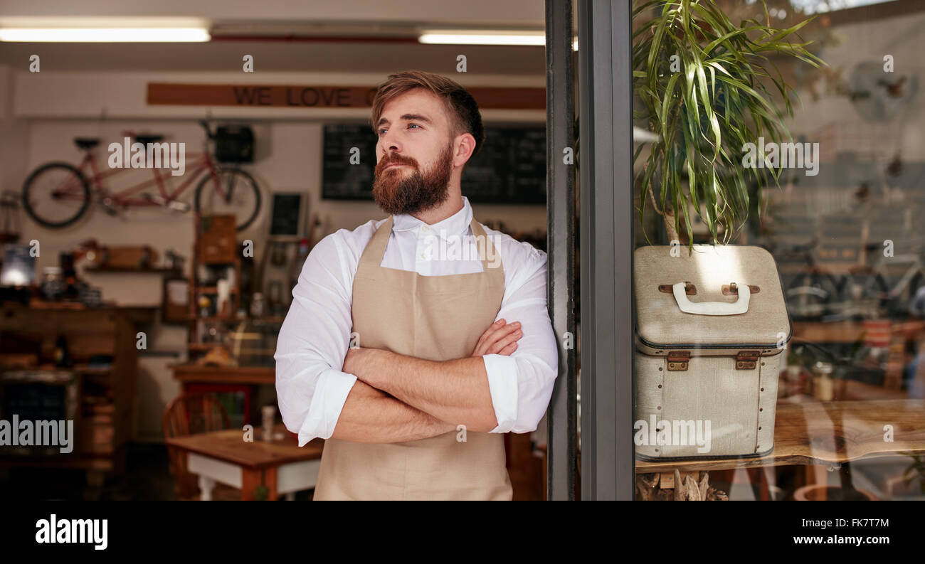 Shot of young man with is arms crossed and looking away while standing at the door of cafe. Barista standing at doorway of a res Stock Photo