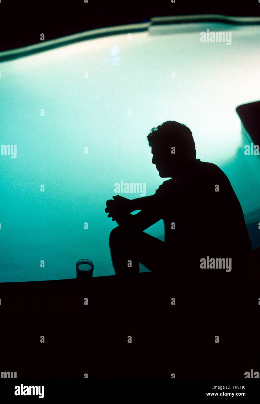 Silhouette of adult male seated by swimming pool with drink Stock Photo