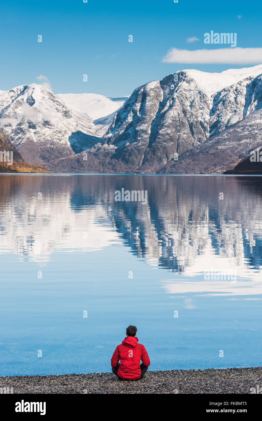 A man in a red jacket sits looking out over Aurlandsfjord in Norway, from Flåm Stock Photo