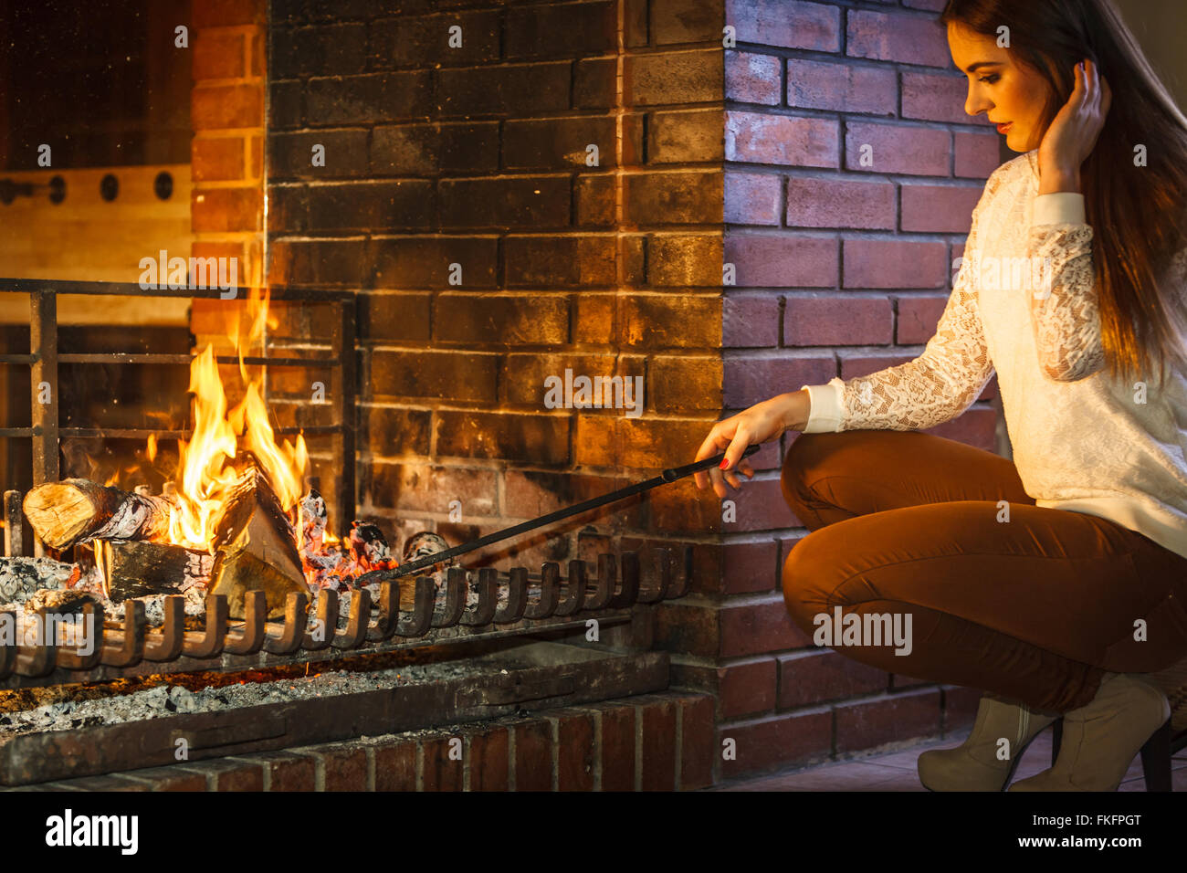 Woman with fire iron poker at fireplace. Young girl heating warming up and relaxing. Winter at home. Stock Photo