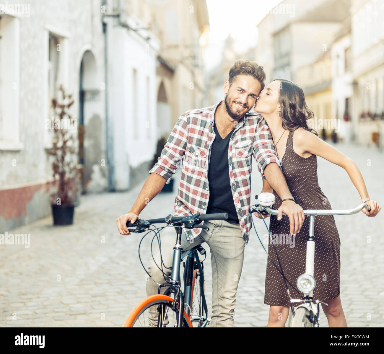 handsome young man with blue eyes stops cycling to be kissed on the cheek by his beautiful partner dressed in brown dress with p Stock Photo