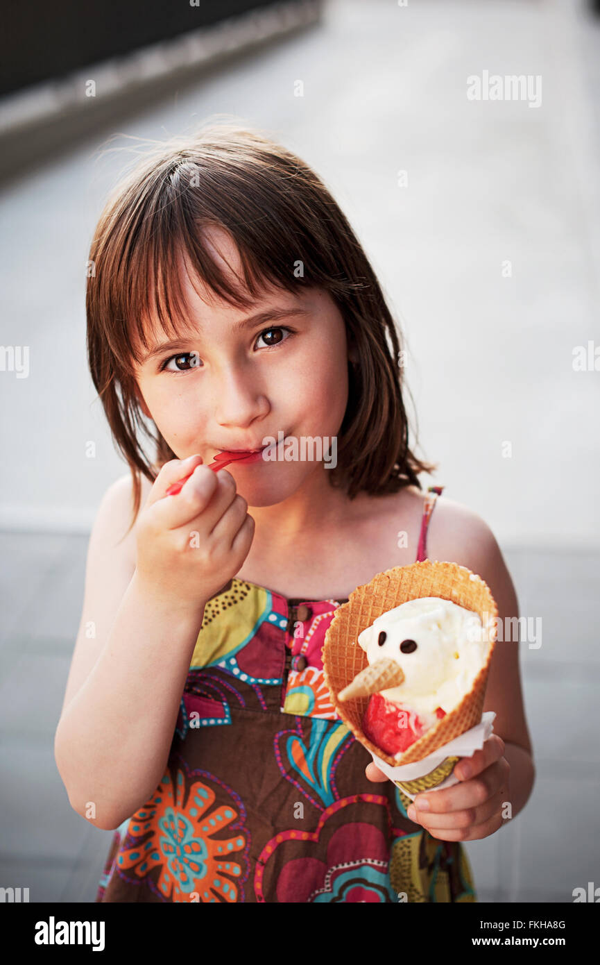 Cute girl eating ice-cream  outside in hot summer day. An ice-cream is made like a snowman with eyes and nose. Stock Photo