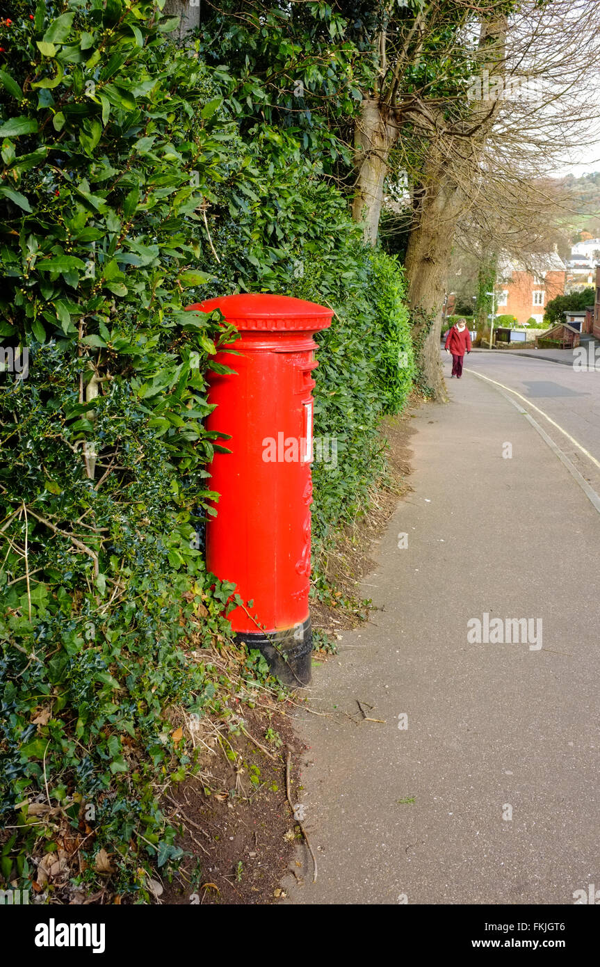 Sidmouth, Devon. Old style red pillar box with female pedestrian walking towards it. Stock Photo