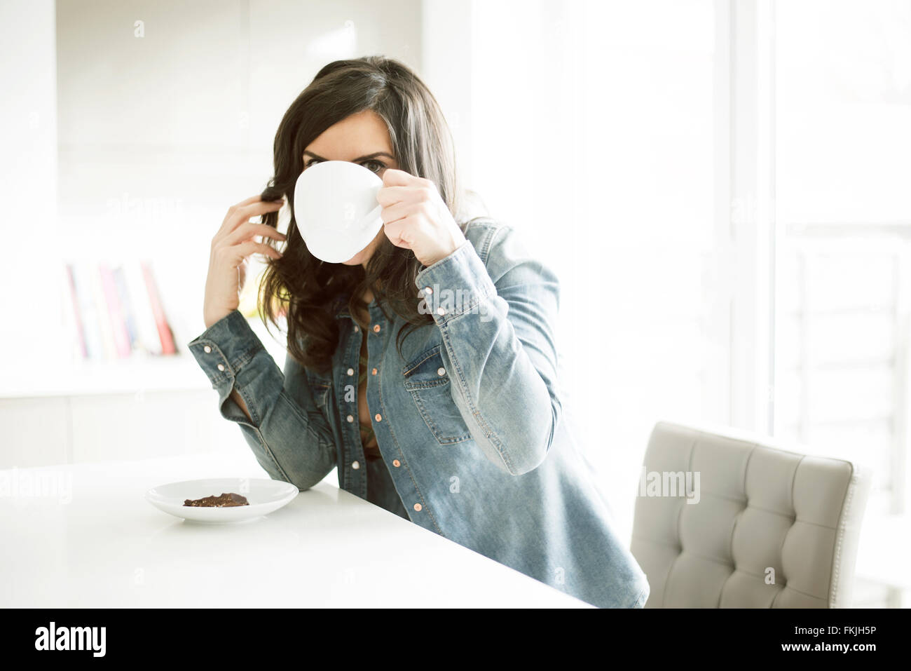 Pretty lady in her 30's drinking coffee in her kitchen wearing blue denim shirt on her teabreak Stock Photo