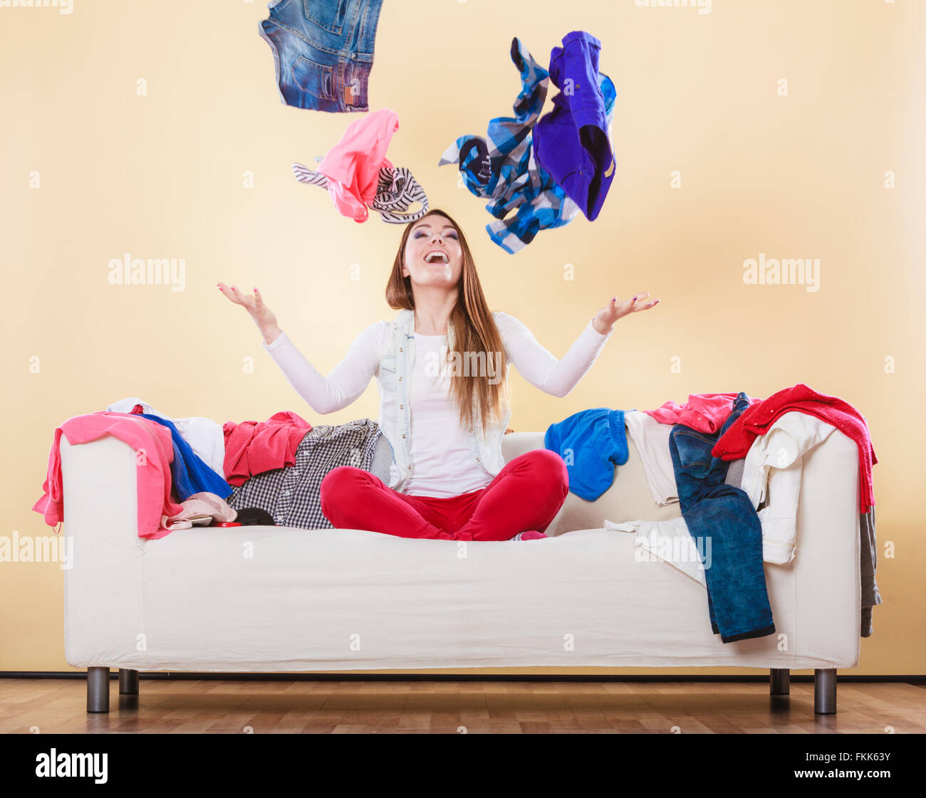Happy woman sitting on sofa couch in messy living room throwing clothes. Young girl surrounded by many stack of clothing. Stock Photo