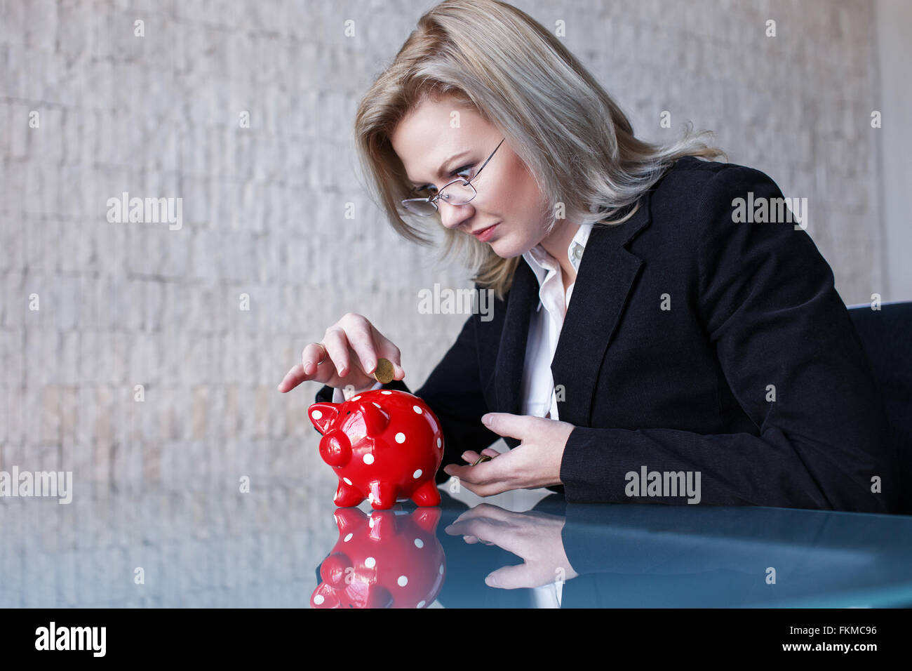Businesswoman in glasses put coins into red dotted piggy bank, saving Stock Photo