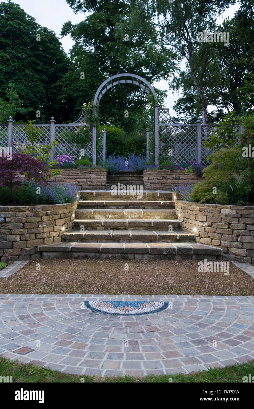 Mosaic art, paving, flowering plants, steps and trellis arch at dusk - designed, landscaped, garden, Burley-in-Wharfedale, Yorkshire, England. Stock Photo