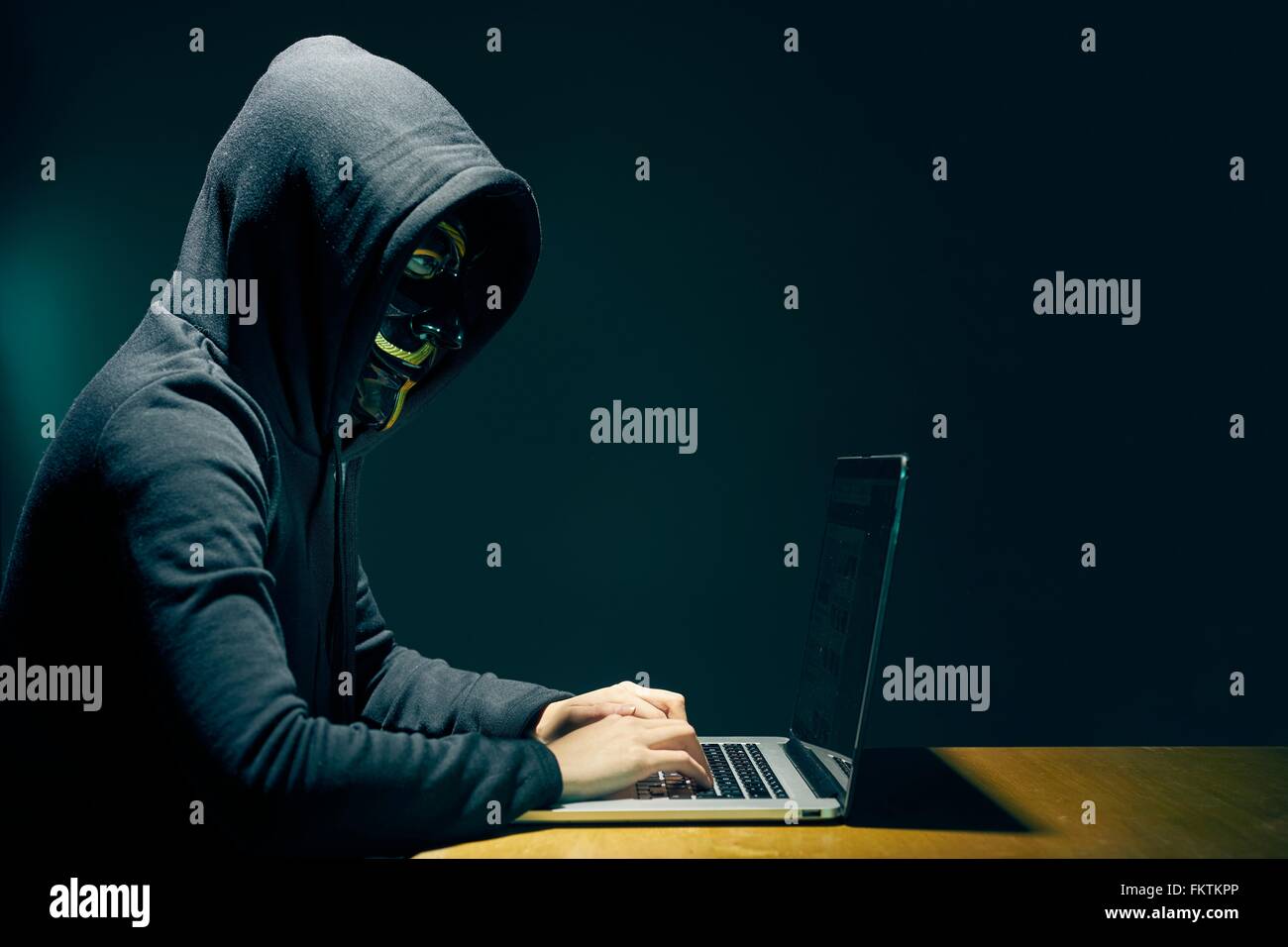 Side view   person wearing hooded top and Guy Fawkes face mask using laptop Stock Photo