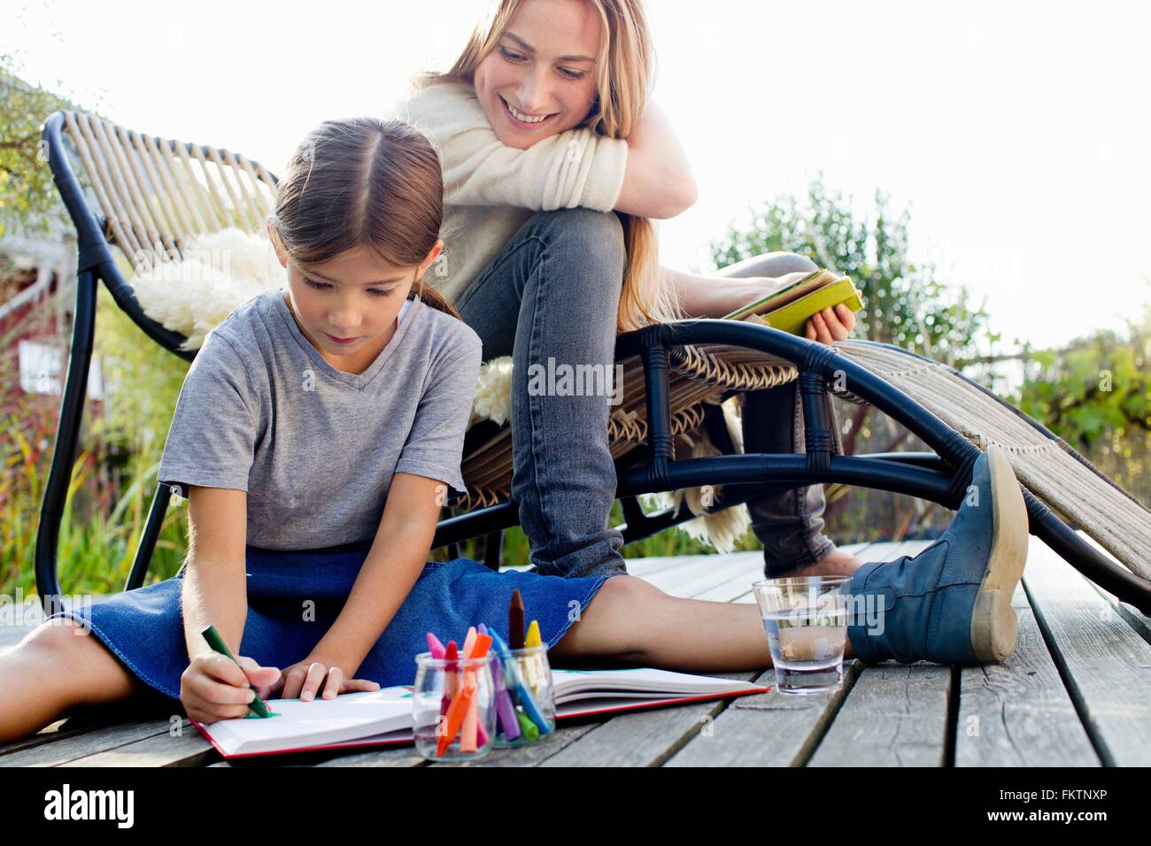 Girl drawing on decking with mother sitting close by, smiling Stock Photo