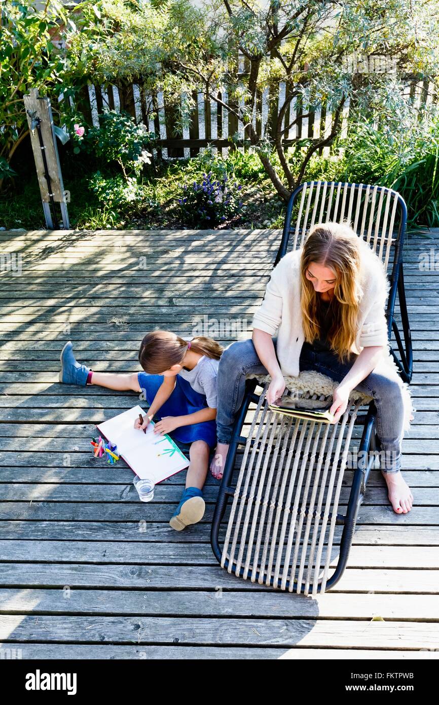 Mother using tablet, daughter drawing on wooden decking Stock Photo
