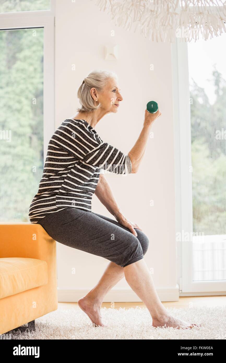 Senior woman sitting on sofa exercising with dumbbell Stock Photo