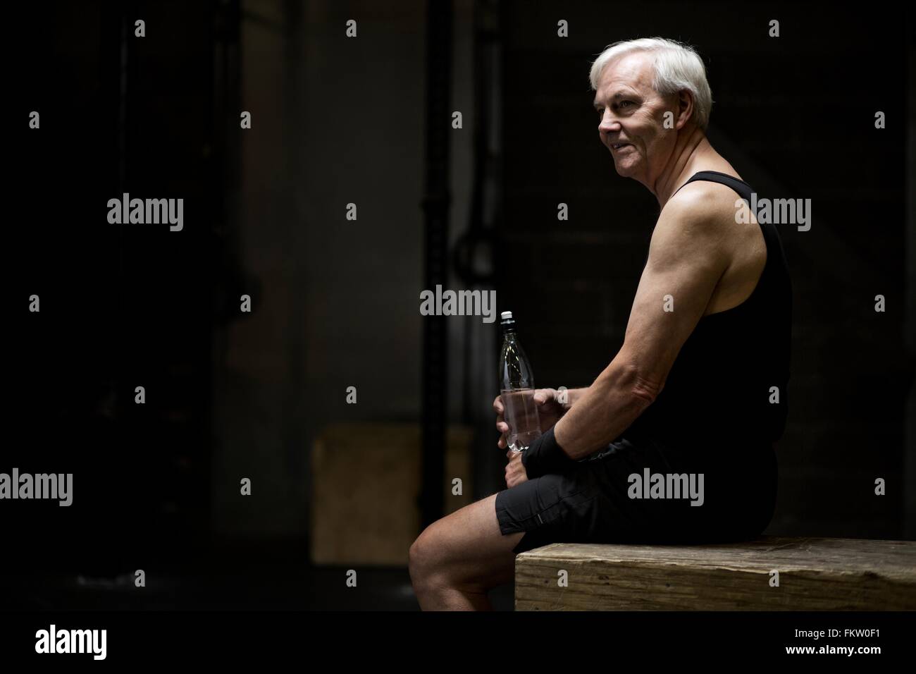 Senior man sitting with water bottle in dark gym Stock Photo