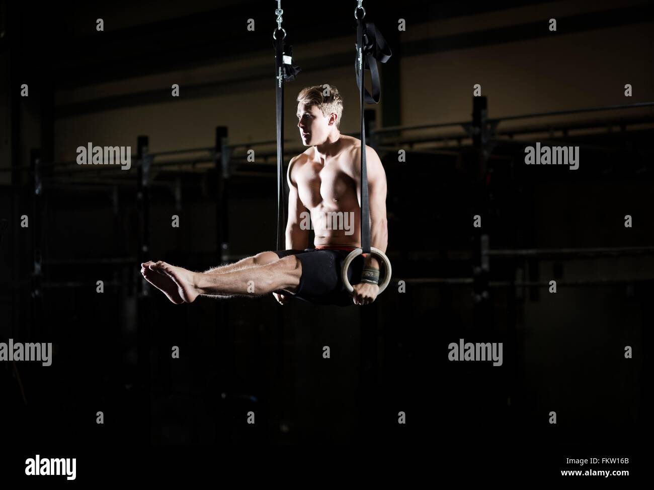 Bare chested young man poised on gym rings in dark gym Stock Photo