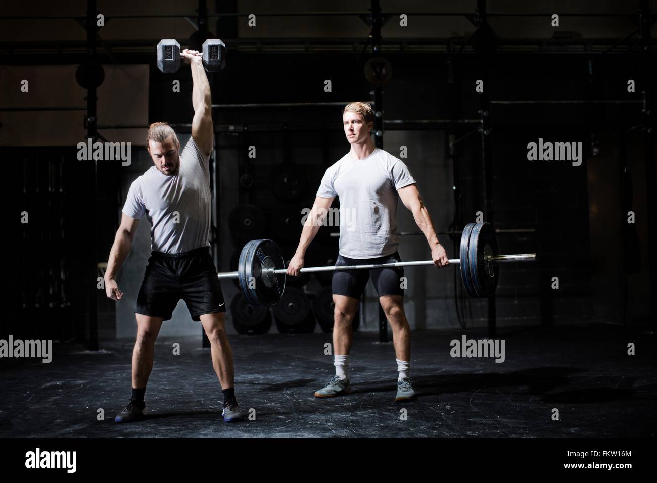 Two young men weightlifting with dumbbell and barbell in dark gym Stock Photo