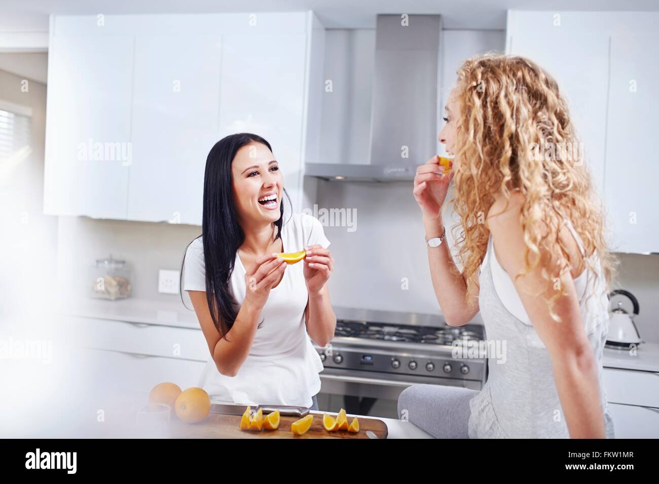 Two young female friends eating breakfast oranges at kitchen counter Stock Photo