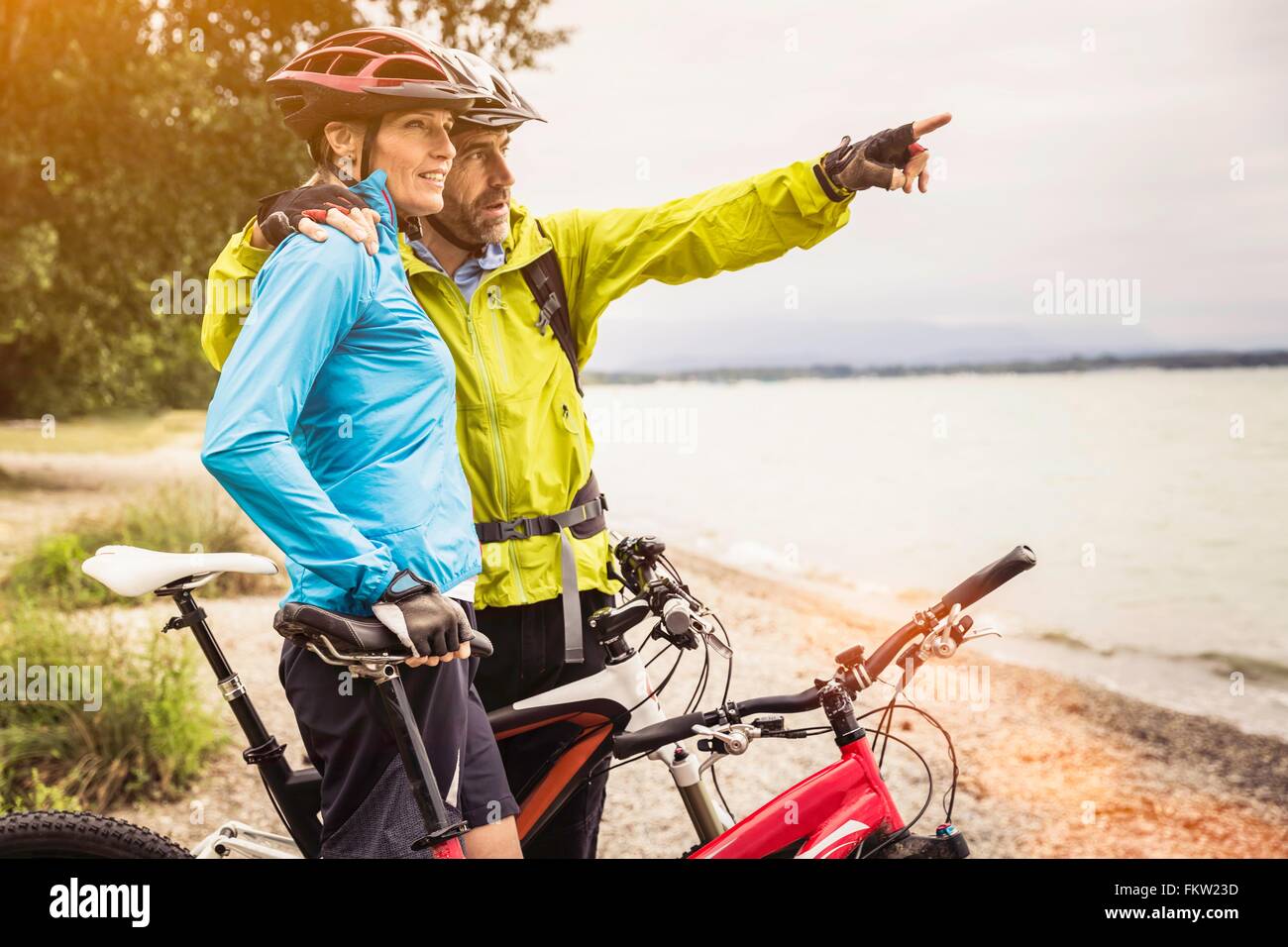 Mature mountain biking couple looking out and pointing from lakeside Stock Photo