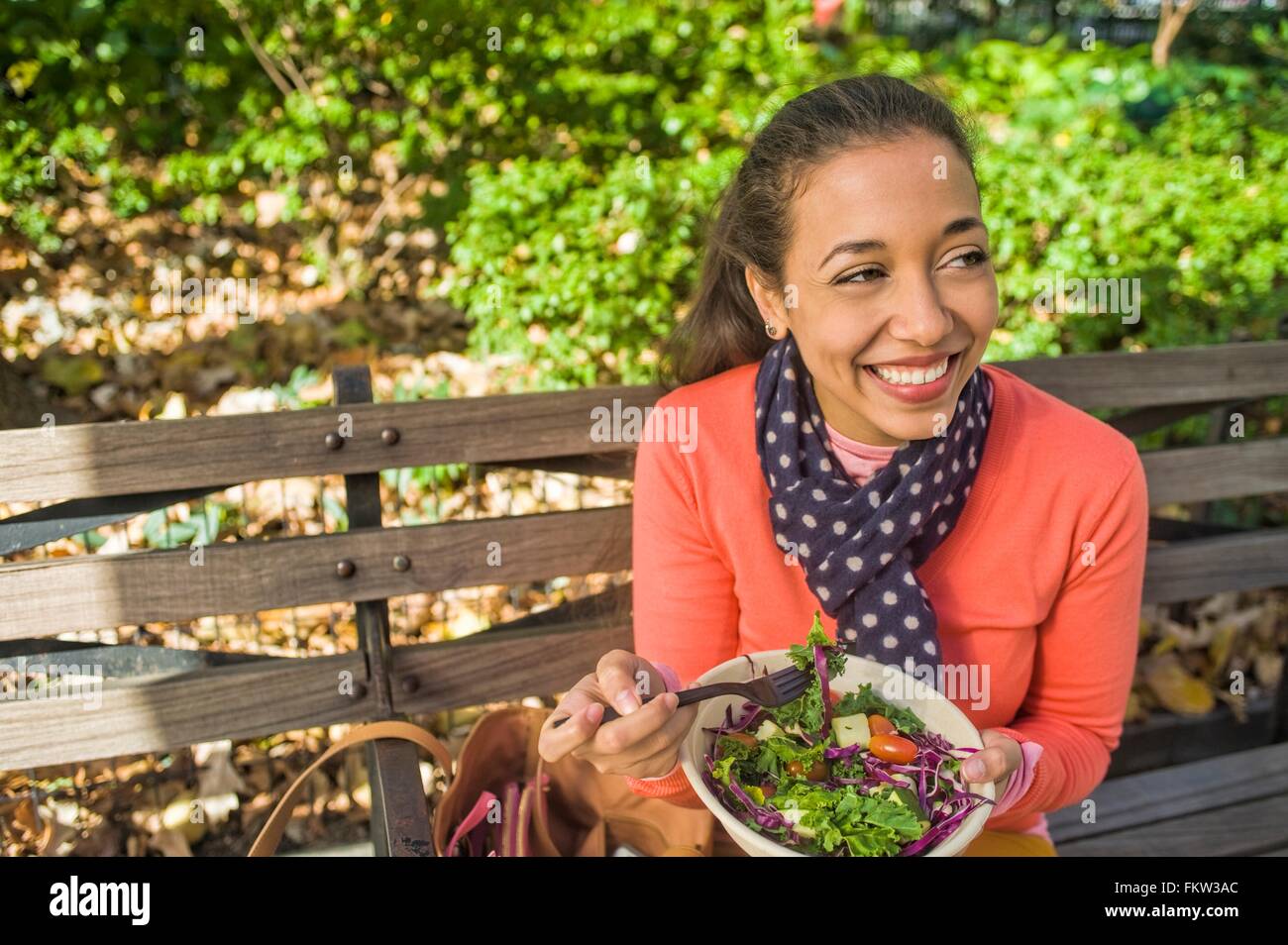 Young woman sitting on park bench eating lunch Stock Photo