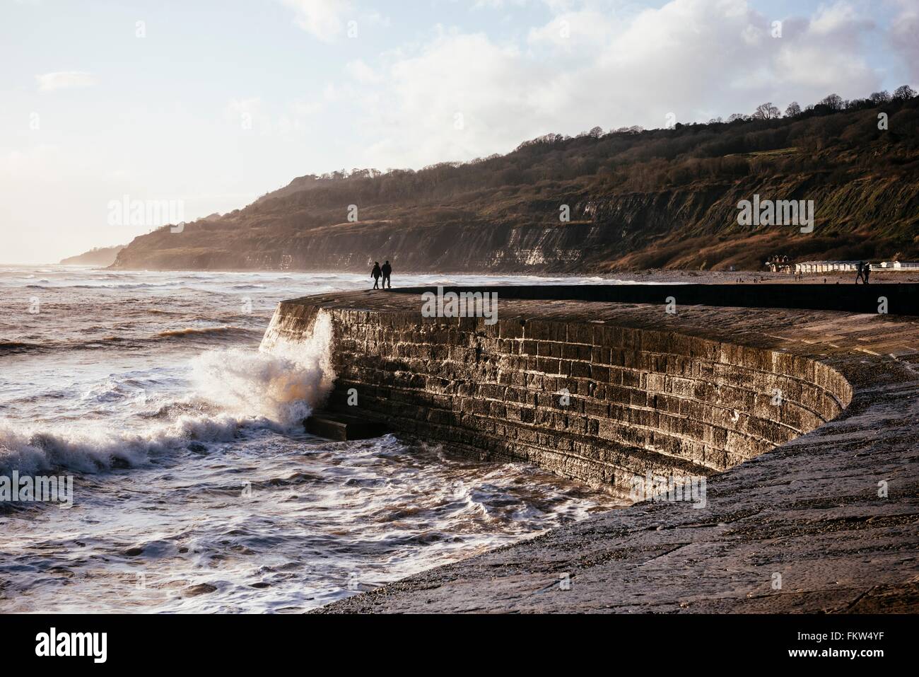 Two people looking out at stormy sea from 'The Cobb' harbour wall, Lyme Regis, Dorset, England Stock Photo