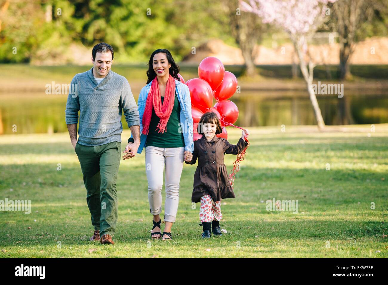 Young couple and daughter with bunch of red balloons strolling in park ...