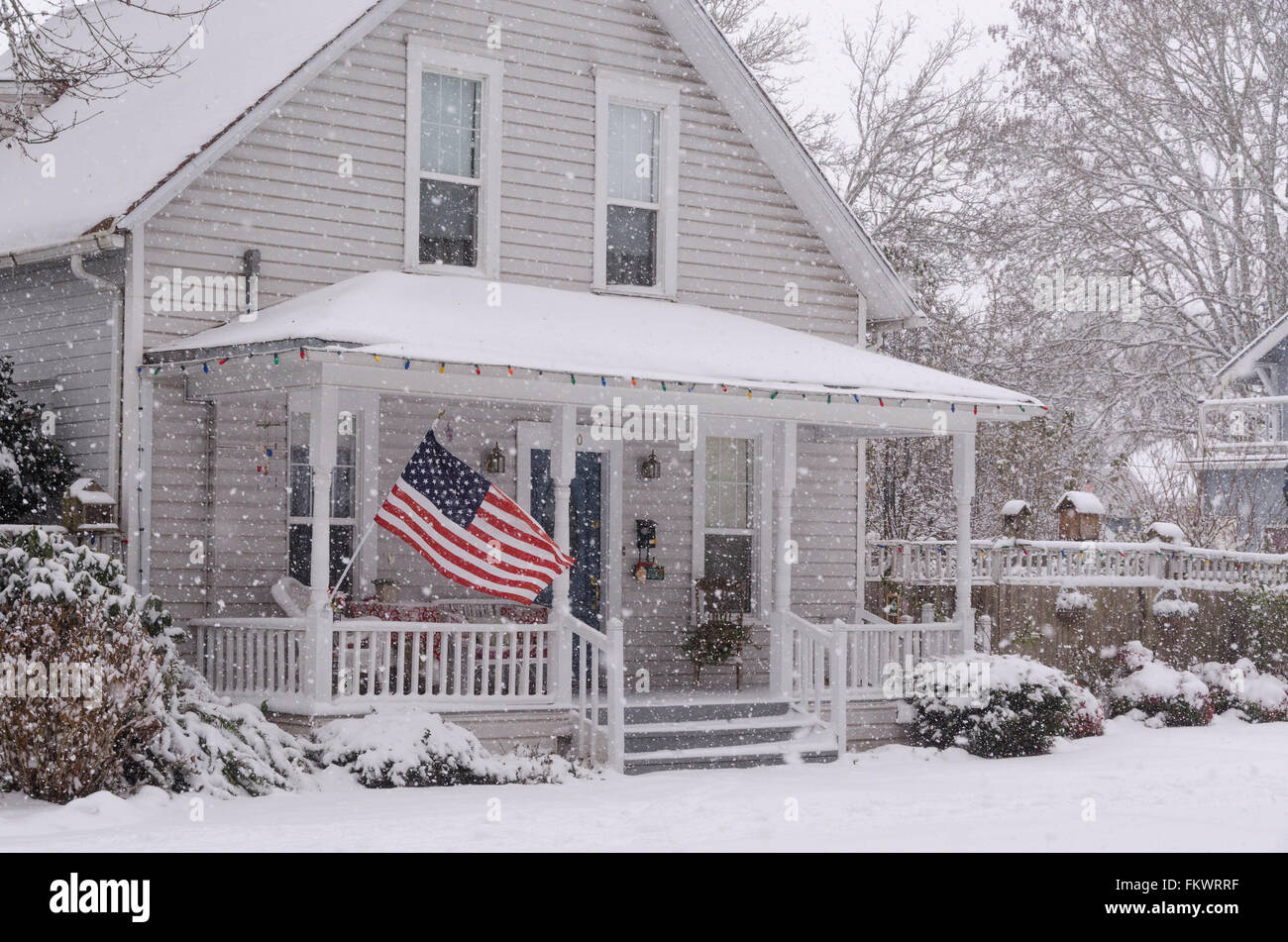 An American flag flies proudly on the porch of an 80 year old house as large snowflakes drift down on the snow-covered ground. Stock Photo