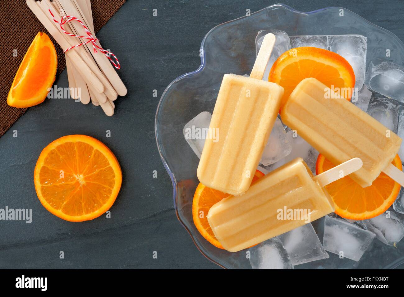 Frozen orange yogurt popsicles in an ice filled bowl with fresh fruit slices against a slate background Stock Photo