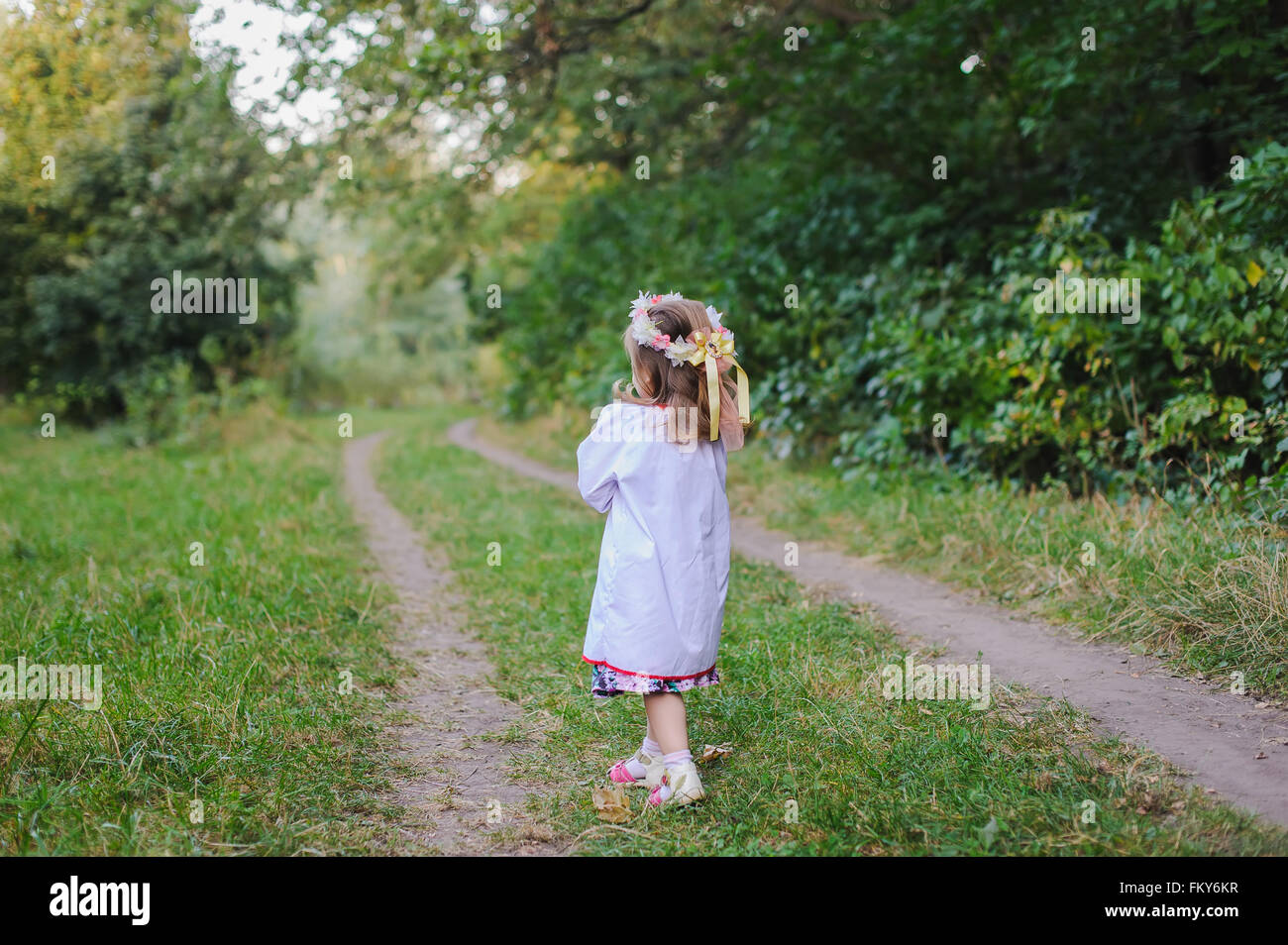 cute little blonde girl in a white dress walking down the path in the forest, the view from the back Stock Photo