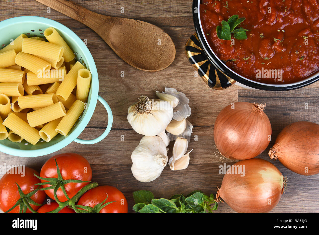 Italian meal ingredients on a wood kitchen table. A pot of sauce, colander of rigatoni, tomatoes, garlic, oregano, and onions se Stock Photo