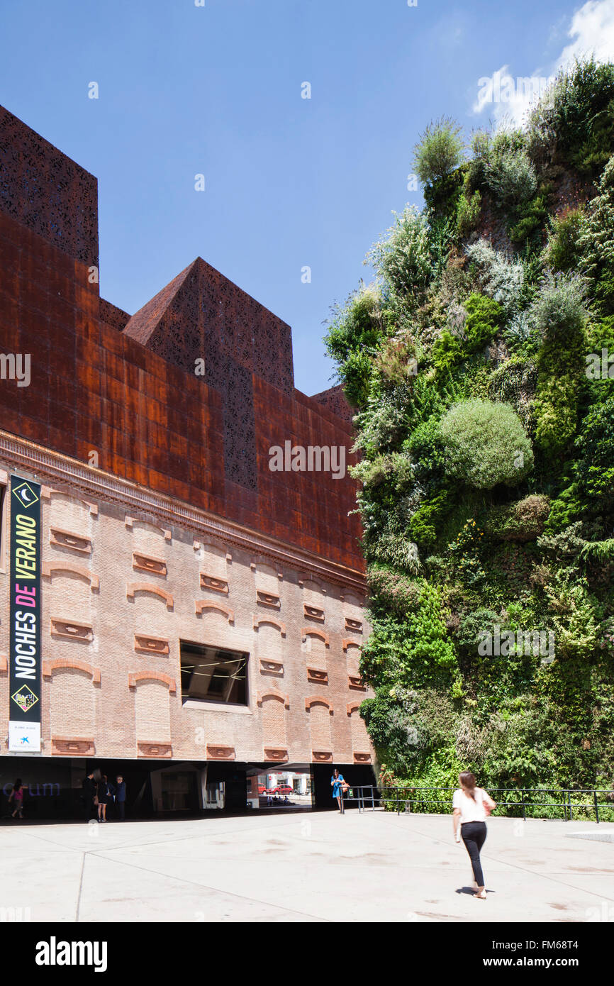 A side view of a building called The Caixa Forum, in Madrid, with the unusual design, as ladies passes between it and a wall of plants next door. Stock Photo