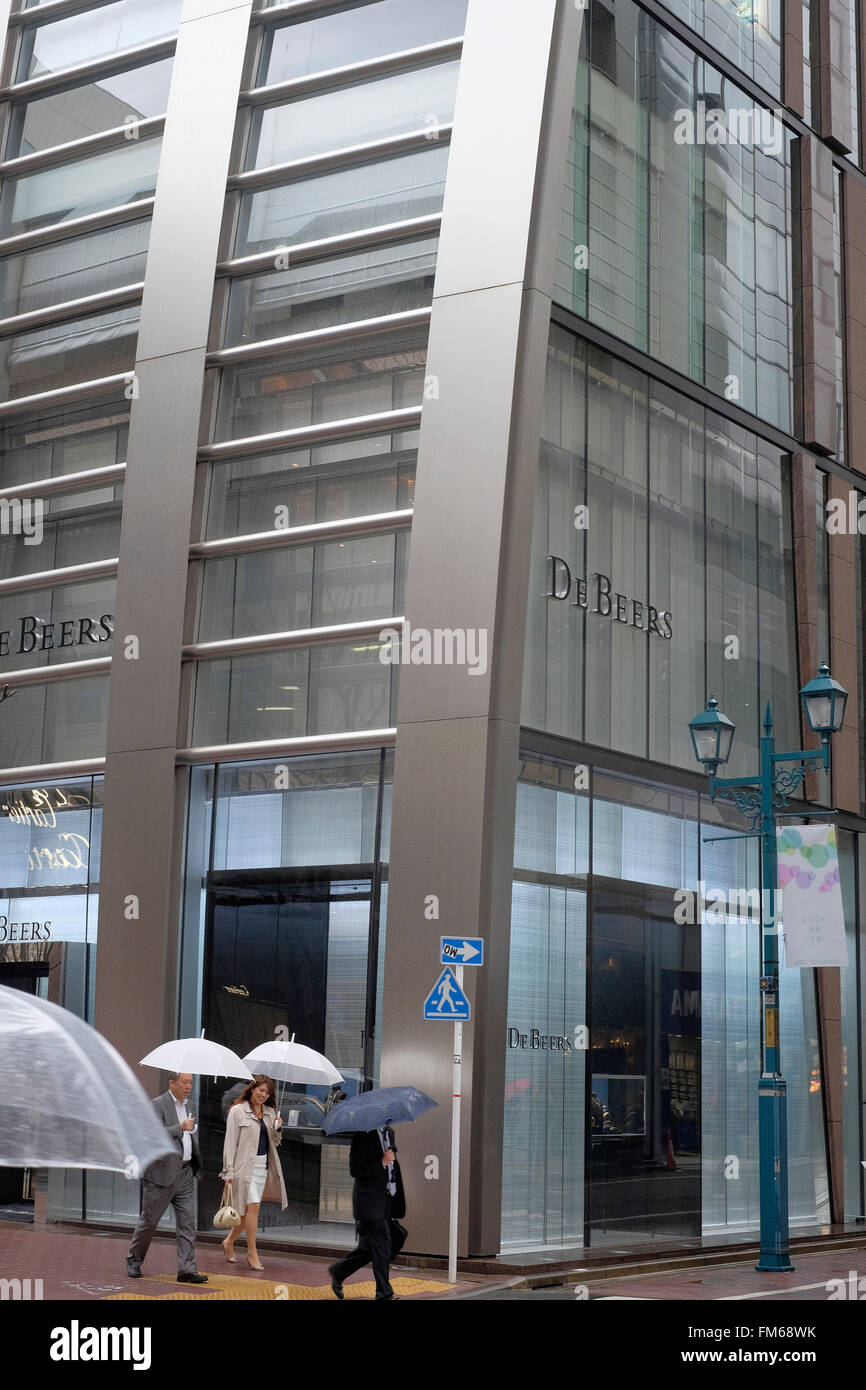 An exterior view of the bottom section of an ultra modern building called the De Beers building in Tokyo, with two men and a woman walking past with raised umbrellas. Stock Photo