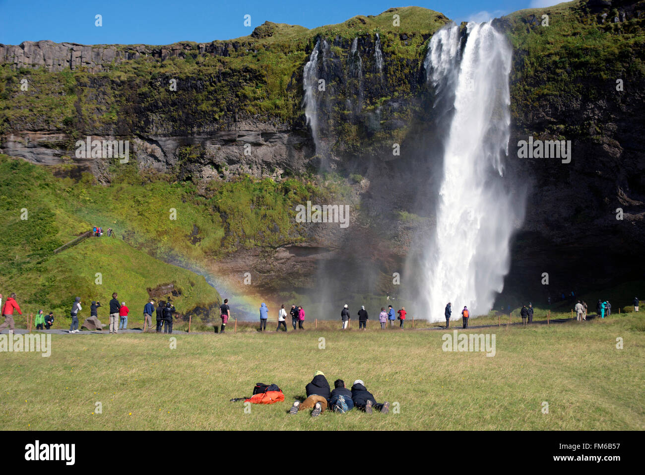 A stunning landscape of a large waterfall called Seljalandsfoss Waterfall , in Iceland, surrounded by fields and mountains, and crowds, and a rainbow. Stock Photo