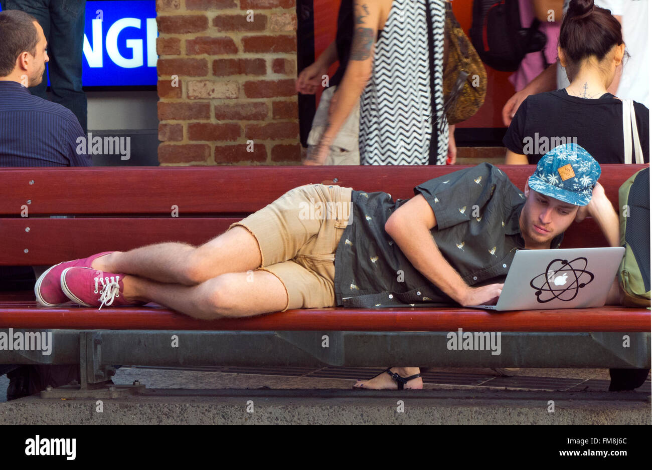 youth in baseball cap laying down on bench using laptop in busy brisbane city street queensland, australia Stock Photo
